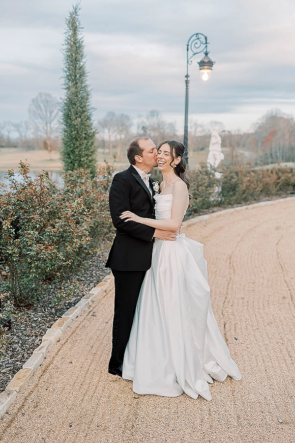 Couple portrait of bride and groom embrace as he kisses her cheek, her strapless gown and lamppost along a garden gravel path