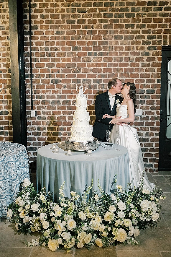 Wedding cake cutting as bride and groom kissing beside a tiered white cake with candles and champagne flutes against a brick wall