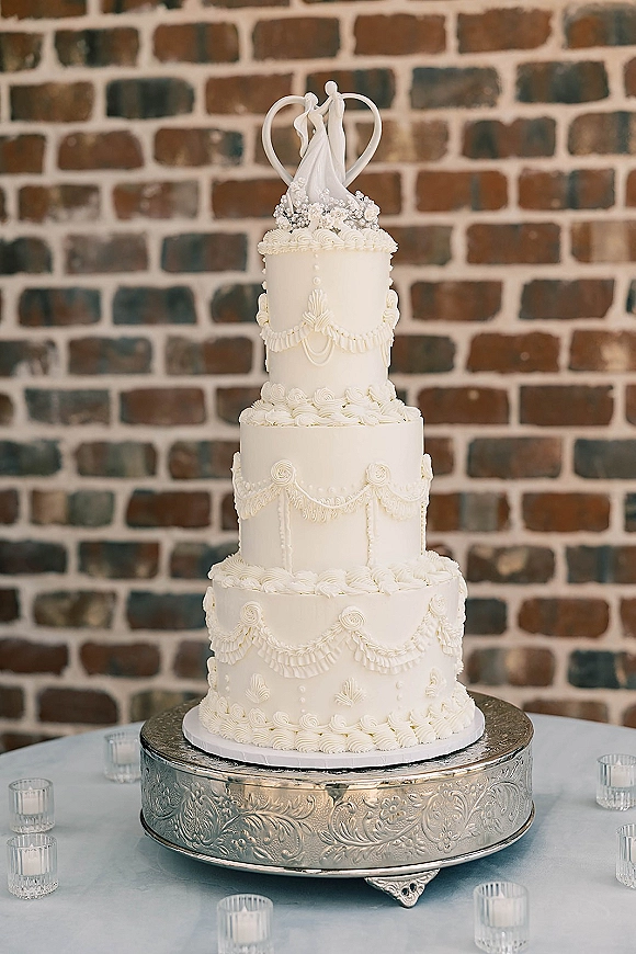 Wedding cake with piped buttercream on a silver stand, topped with figurines and surrounded by votive candles against a brick wall