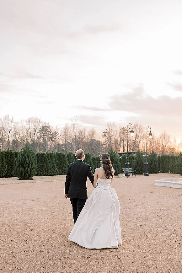 Couple portrait of bride and groom walking away hand in hand, strapless gown and tuxedo in a sunset estate courtyard with lampposts