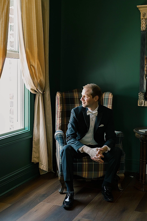 Groom portrait in a black tuxedo with white bow tie, sitting in a plaid armchair and looking out a window by a green wall