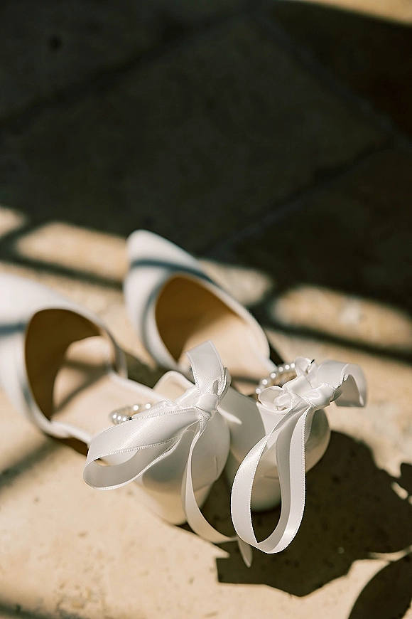 Bridal shoes with white bow heels, pearl detail and pointed toes resting on stone steps, sunlight casting soft shadows