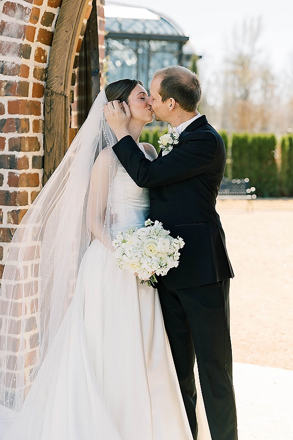 Wedding kiss portrait of bride and groom kissing, her veil flowing as she holds a white bouquet with greenery beside a brick wall doorway