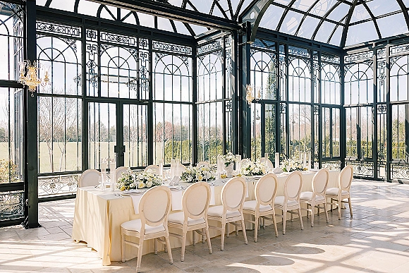 Reception tablescape with long head table decor, ivory linens, white florals, greenery, and candles under chandeliers in a glass conservatory