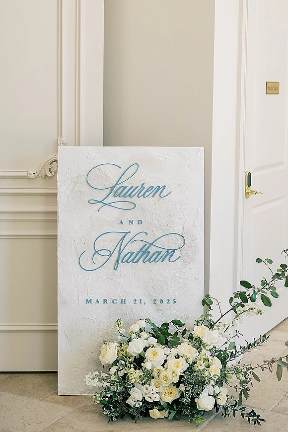 Wedding welcome sign with custom wedding sign calligraphy on a textured white plaster board, leaning beside white roses and greenery indoors