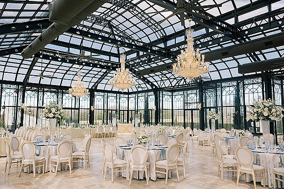 Reception space in a glass conservatory reception with crystal chandeliers, white linen round tables, tall florals, and windowed views