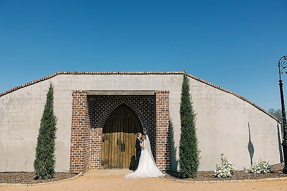 Wedding couple portrait of bride and groom kissing in front of wooden doors under a brick archway, bridal veil flowing on gravel walkway