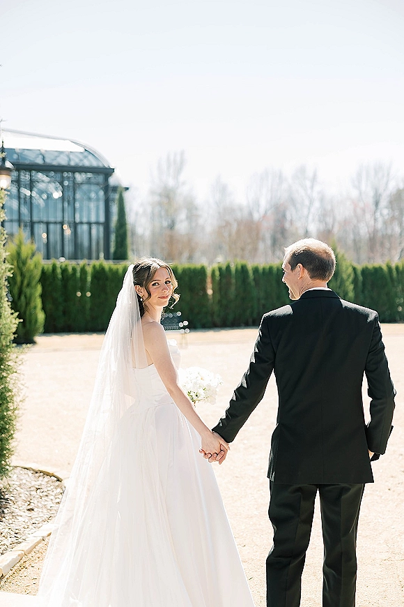 Couple portrait of bride and groom walking away holding hands, bride glancing back, cathedral veil flowing on a garden path by a greenhouse