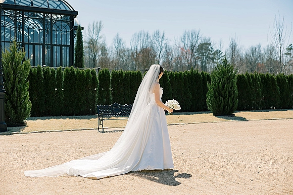 Bridal portrait of a bride holding white bouquet with a cathedral veil and long train in a glass conservatory courtyard under blue sky