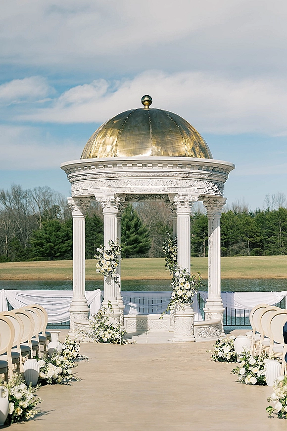 Ceremony setup for an outdoor wedding ceremony with aisle floral arrangements, white draping and chairs on a lakefront lawn under trees