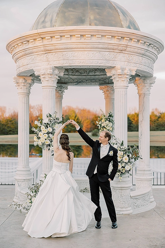 Couple portrait of bride and groom twirl under a white gazebo, her strapless gown sweeping as sunset glows over the lakeside lawn