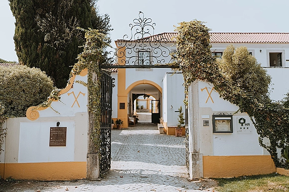 Wedding venue exterior with a wrought iron gate draped in ivy, leading to a white stucco arched entryway with lantern and potted plants.