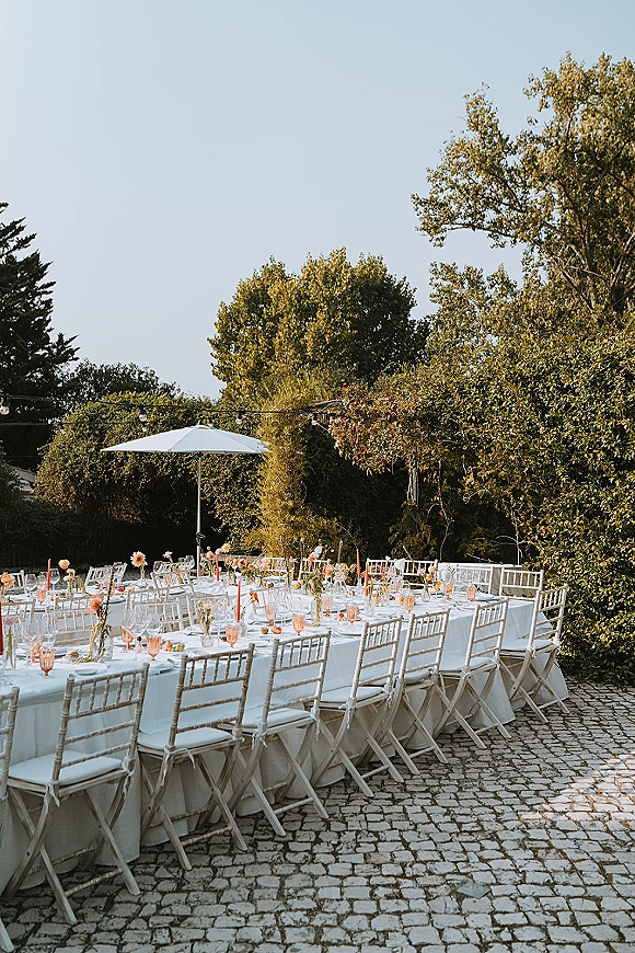 Reception tablescape with long banquet tables in white linens, pink taper candles, bud vases, and string lights on a cobblestone patio