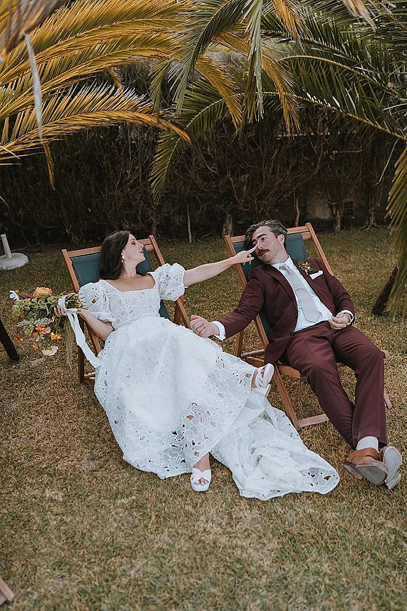 Couple portrait of bride and groom lounging on deck chairs, her bouquet and lace puff sleeves visible on a palm-lined lawn