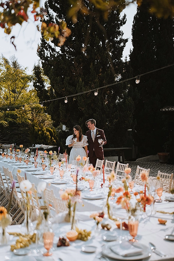 Reception tablescape with white tablecloth, peach goblets, bud vases and taper candles on a long banquet table under garden string lights, bride and groom toasting