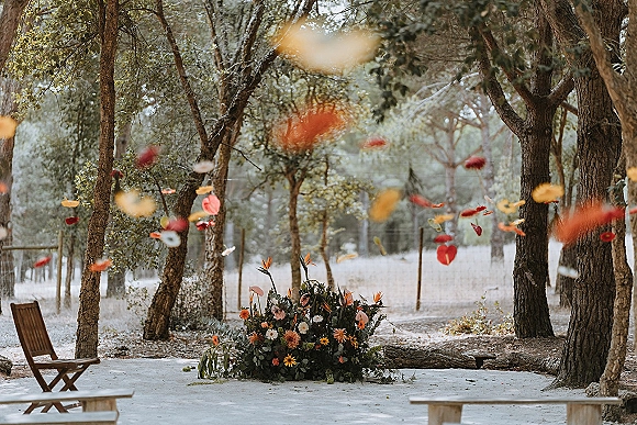 Ceremony aisle decor with ground floral arrangement and scattered petals, framed by wooden chairs and benches in a forest clearing