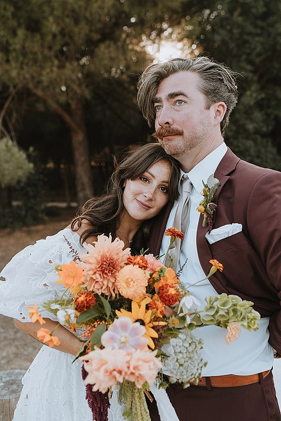 Couple portrait of bride leaning on groom, holding an orange wedding bouquet, in warm sunlight with trees and outdoor greenery behind
