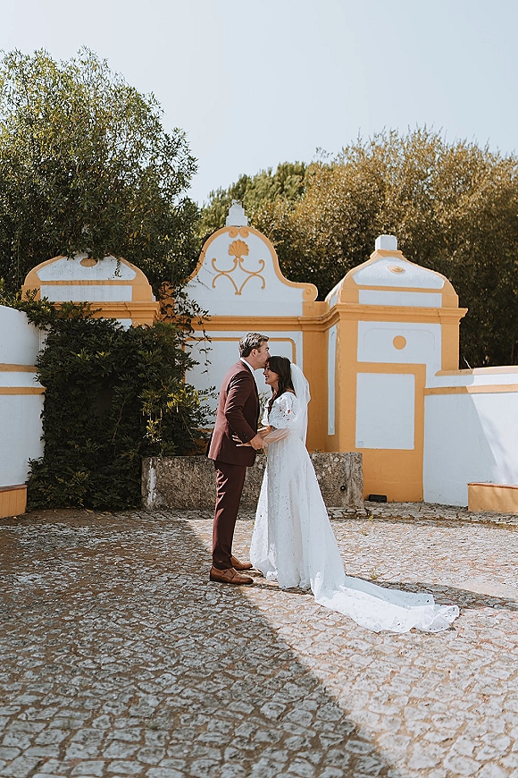 Couple portrait of bride and groom embrace in a forehead kiss, her lace-sleeve dress and veil against a white and yellow wall courtyard