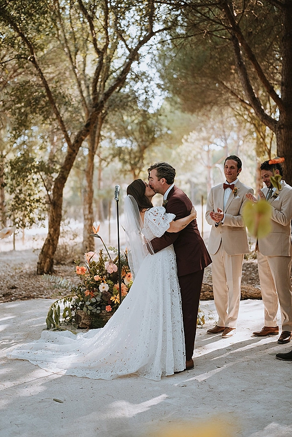 Wedding kiss at an outdoor wedding ceremony as the bride in a lace dress and veil kisses the groom in a burgundy suit in a sunlit forest