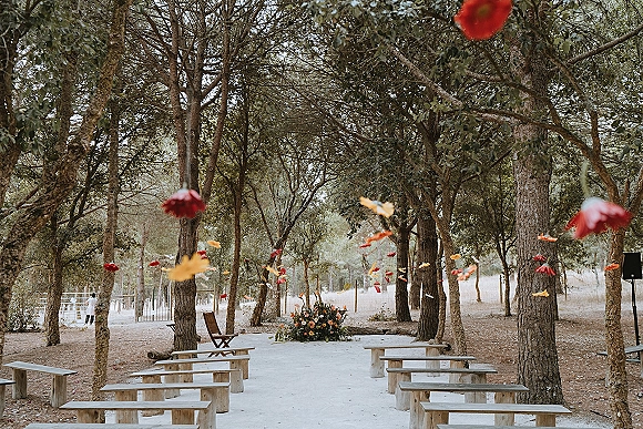 Ceremony setup with wood benches and a white aisle runner, colorful hanging paper flower decor in a pine forest clearing