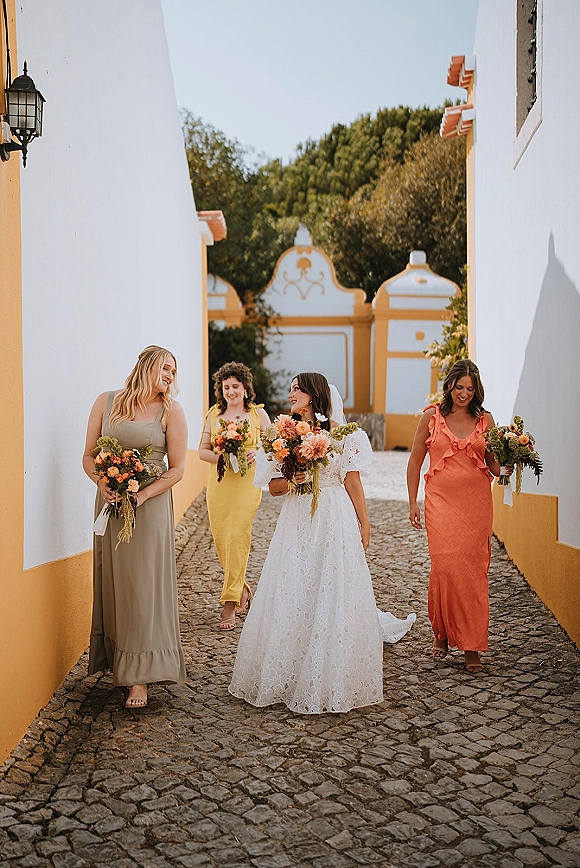 Bride with bridesmaids walking photo on a cobblestone street, holding bouquets, bride in lace gown and veil by white and yellow buildings