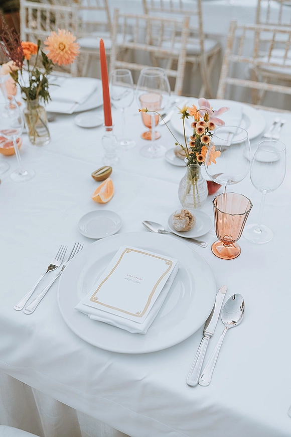Reception tablescape with wedding place setting on a white tablecloth, pink goblets, orange flowers, and taper candle at an outdoor reception area