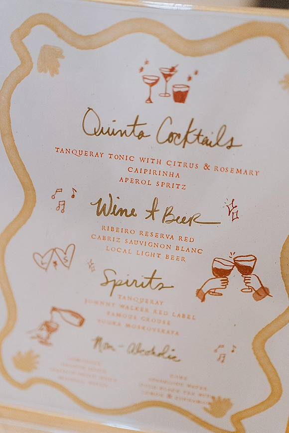 Wedding bar menu with calligraphy lettering and illustrated cocktail icons, framed by a gold border on white paper atop a blurred tabletop