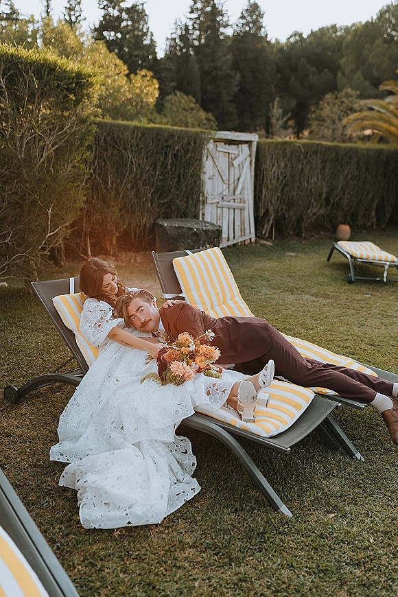 Couple portrait of bride and groom lounging on striped chairs, bride leaning on groom with bouquet on a garden lawn by hedges