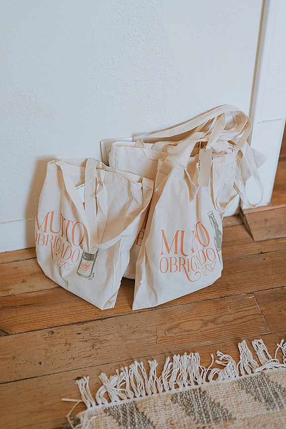 Wedding welcome bags in canvas tote bags with printed lettering and gift tags arranged on a wood floor beside a woven rug