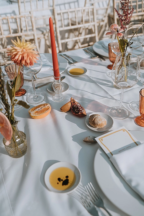 Reception tablescape with a wedding place setting on white tablecloth, taper candles, bud vase florals, amber goblets, and pomegranate accents outdoors