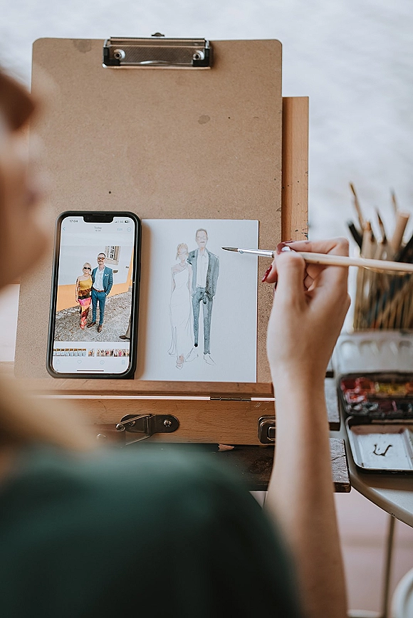 Wedding illustration in progress on watercolor paper, with a clipboard and smartphone reference on an artist desk with paintbrushes and palette