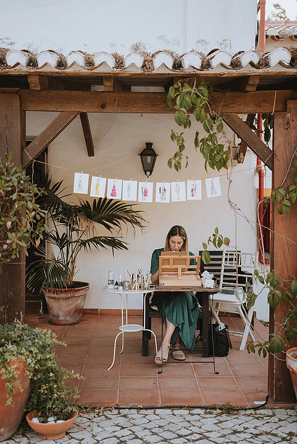 Wedding live painter at a live wedding painter setup with easel, paints and palette, art prints clipped on string in a terracotta courtyard patio