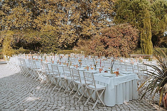 Outdoor reception tablescape with long banquet tables in white linens, pink goblets, bud vases, taper candles and string lights on cobblestone patio