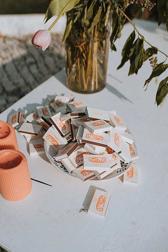 Wedding matchboxes and custom matchbox favors arranged on a ceramic plate with ribbed pillar candles, tulip vase, and sunlit shadows