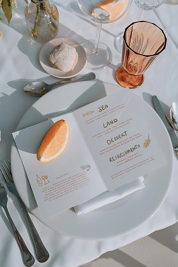 Wedding menu card on a dinner plate with wedding place setting, silver flatware and amber glassware on white linen in sunlit shadows