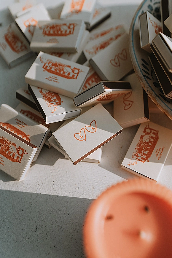 Wedding matchboxes with custom wedding matchboxes sleeves in orange heart illustrations beside a candle and ceramic bowl on a white tabletop