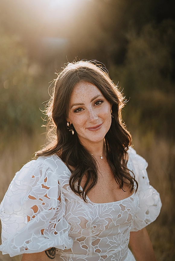 Bridal portrait of a smiling bride in a puff-sleeve lace wedding dress, backlit in tall grass during golden hour sunlight