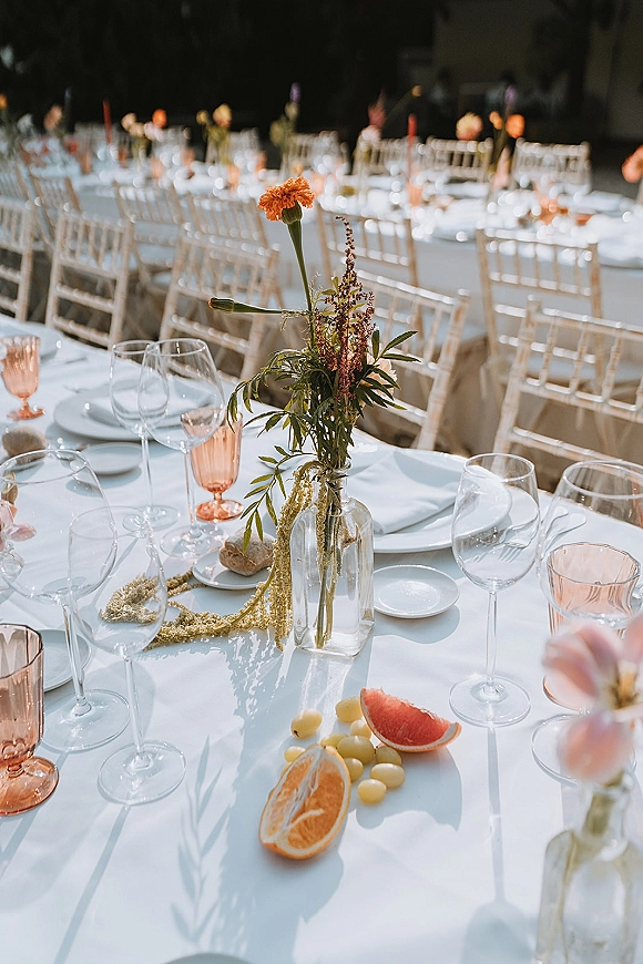 Reception tablescape on a long banquet table wedding with white linen, bud vase orange florals, pink goblets, and citrus fruit place settings