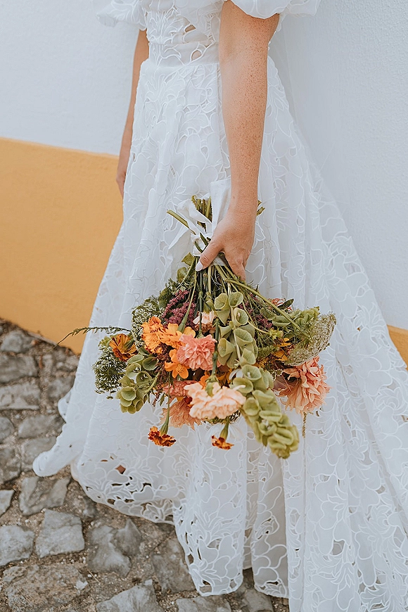 Bridal bouquet with cascading wedding bouquet shape, orange and peach blooms and green hydrangea held against a white lace puff-sleeve dress.