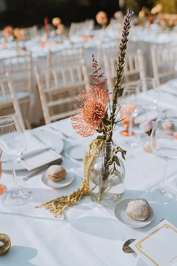 Reception tablescape with wedding bud vase centerpiece featuring orange pincushion flower, dried florals, peach goblets, and white place settings on linen tablecloth