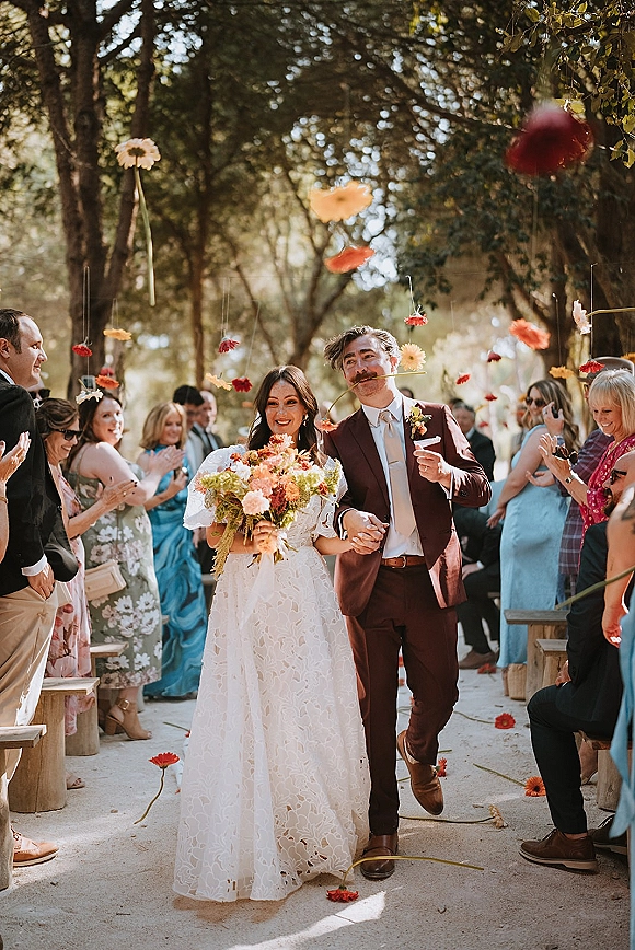 Wedding recessional as bride and groom walk hand in hand down an outdoor aisle while guests cheer and toss flower petals under trees