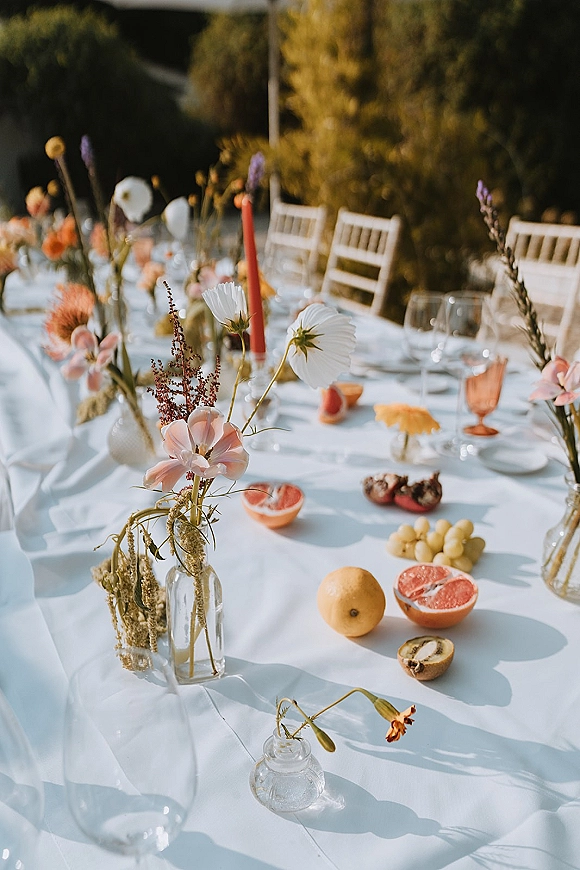 Reception tablescape with long banquet table decor, wildflower bud vases and red taper candles, fruit and amber goblets in a garden setting