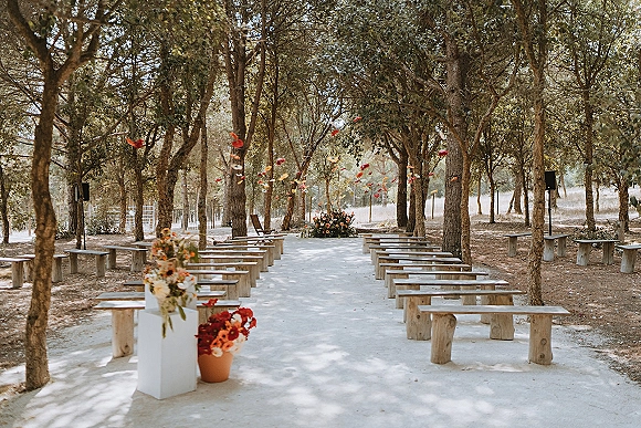 Ceremony setup for an outdoor wedding ceremony with white aisle runner, wood bench seating, and hanging floral plinths in a woodland clearing