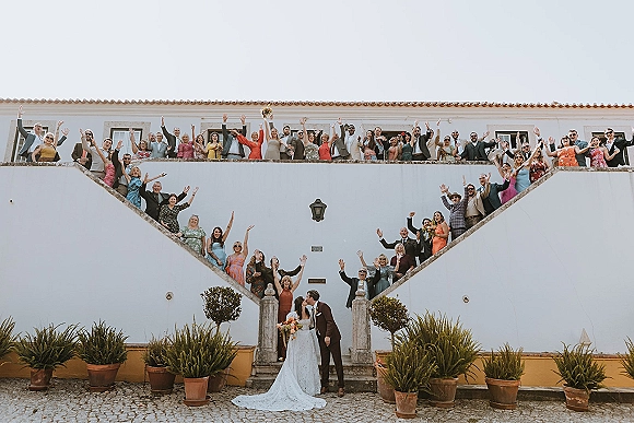 Wedding group photo with guests cheering on an outdoor staircase, bride holding bouquet beside groom in front of white stucco building