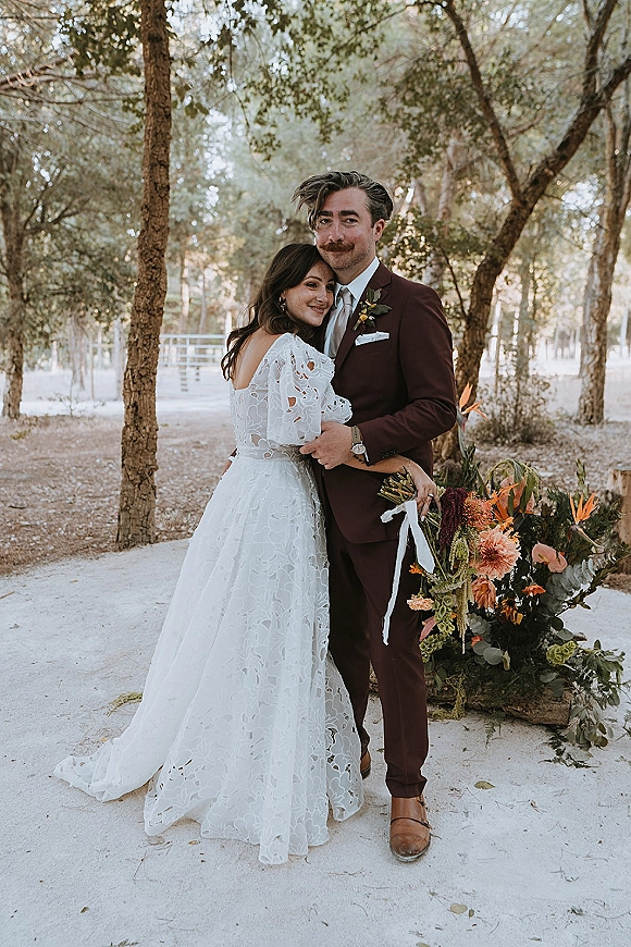 Couple portrait of bride leaning on groom as he holds a rust orange bouquet with ribbon tails on a sandy forest path among trees