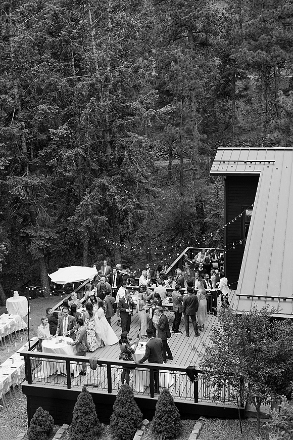 Wedding reception with outdoor wedding reception cocktail tables under string lights on a deck, forest hillside and modern building behind