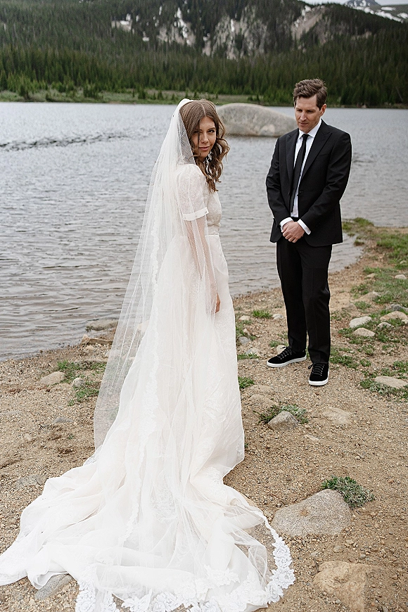 Couple portrait at a lakeside wedding portrait, bride in lace dress with long veil train and groom in suit on rocky shore by mountains