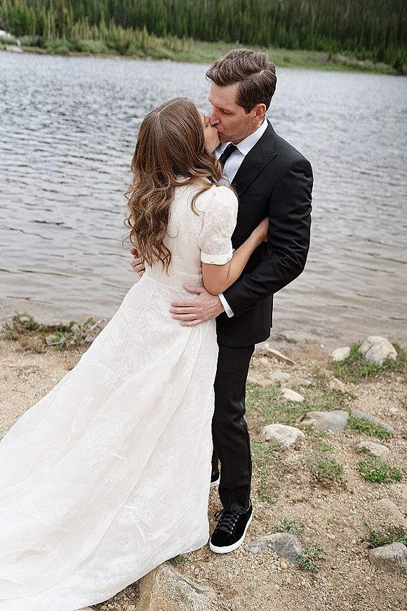 Wedding kiss portrait of bride and groom kissing, her lace short-sleeve gown and his tuxedo with sneakers on a rocky lakeshore by evergreen forest