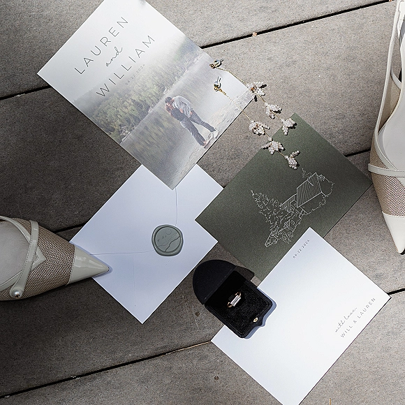 Wedding invitations and wedding invitation suite arranged on a wood deck with envelopes, gray wax seal, rings, pearl hair vine and shoes