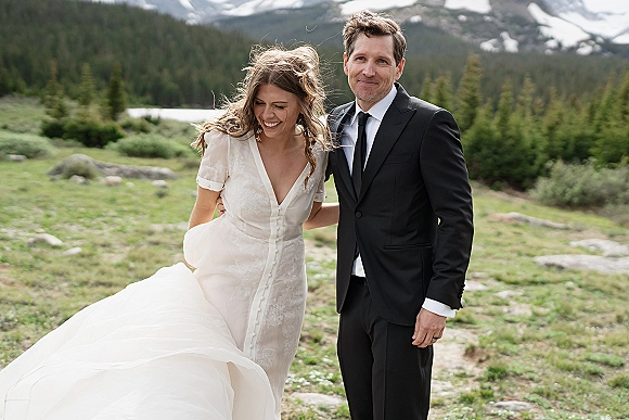 Couple portrait of bride and groom laughing, groom arm around her as she holds her dress train in an alpine meadow by a lake